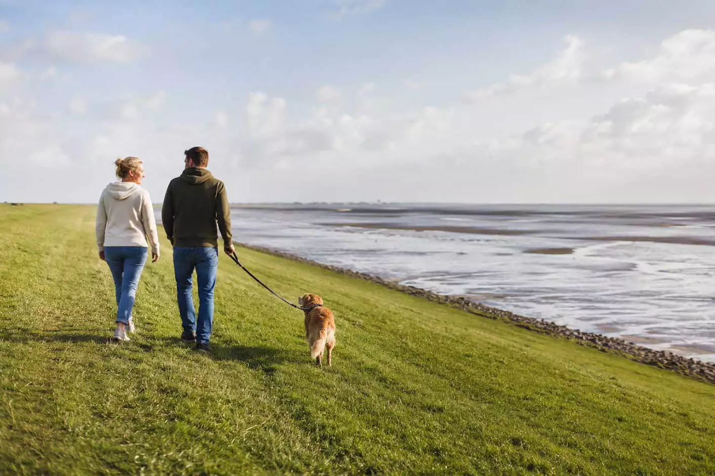 Paar mit angeleintem Hund beim Spaziergang auf dem grünen Deich mit Blick über das Wattenmeer auf Nordstrand